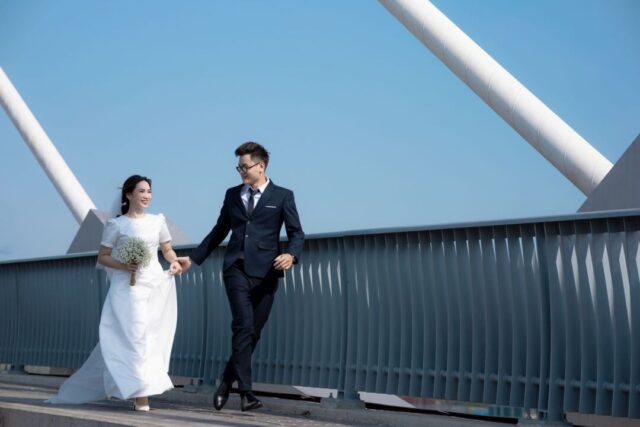 Smiling couple in simple modern attire holding a small bouquet on the marble steps of a city courthouse, candid...