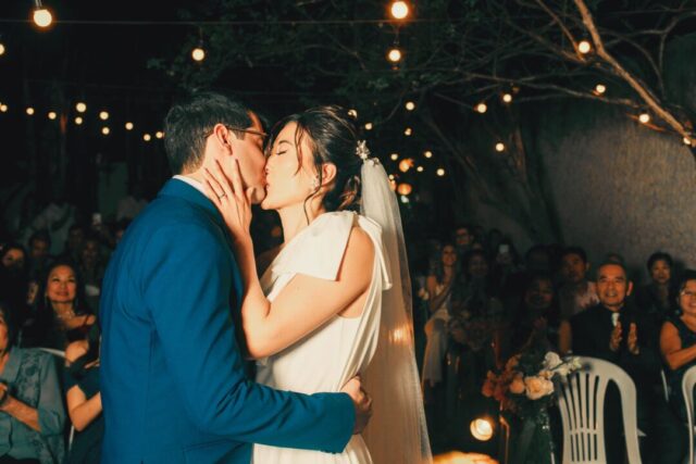 Joyful couple sharing their first kiss at an intimate micro wedding in a sunlit garden, surrounded by 25 elegantly...