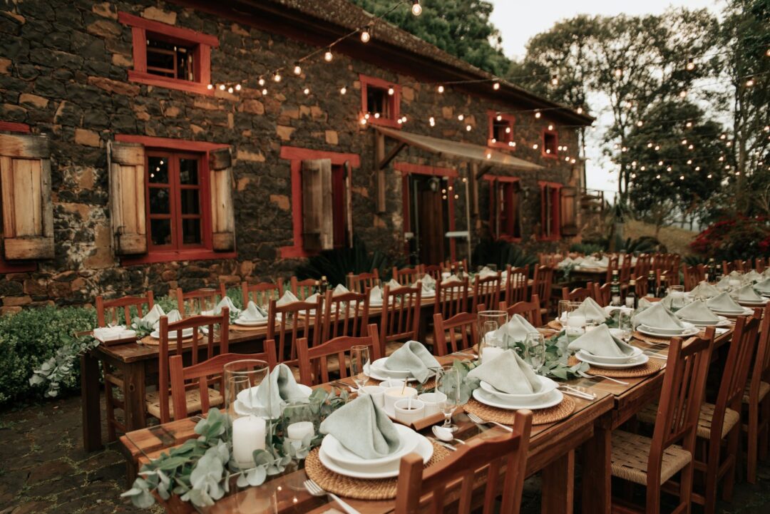 Backyard rehearsal dinner at dusk, long farm tables set with white linens and simple wildflower centerpieces, string...