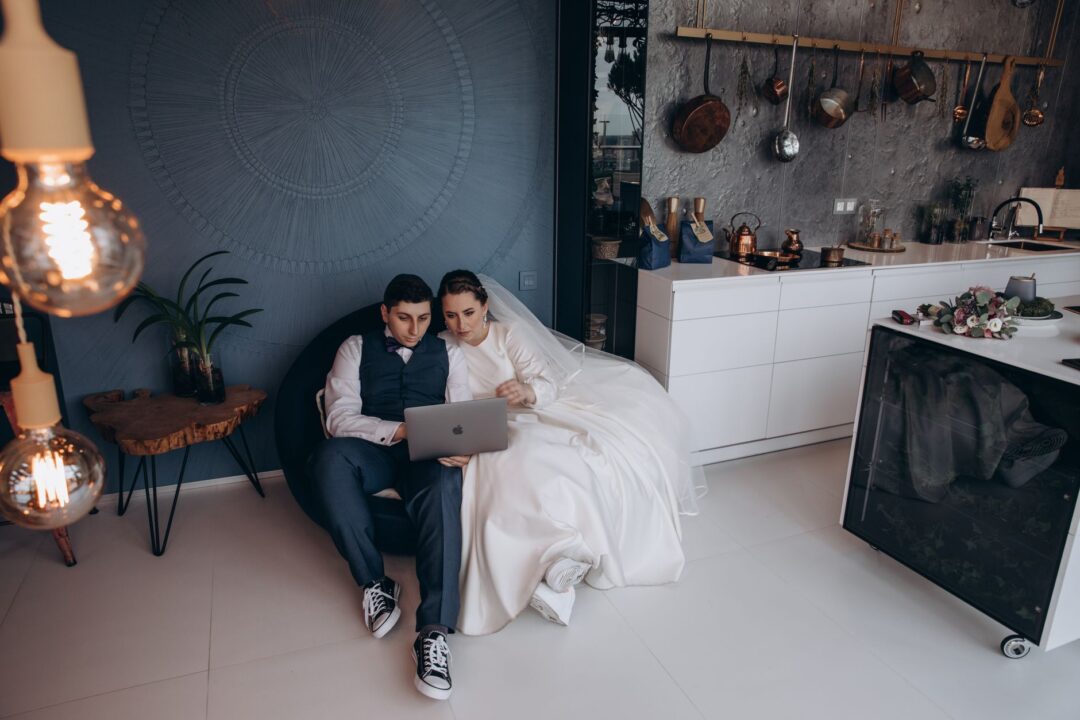 Couple seated together at a kitchen table planning their wedding budget, laptop open to a spreadsheet, handwritten notes...