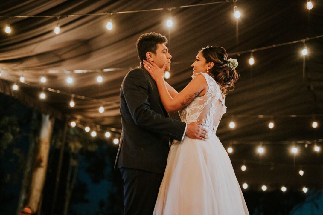 Couple sharing their first dance in a warmly lit rustic barn venue while a live band performs on a small elevated stage...