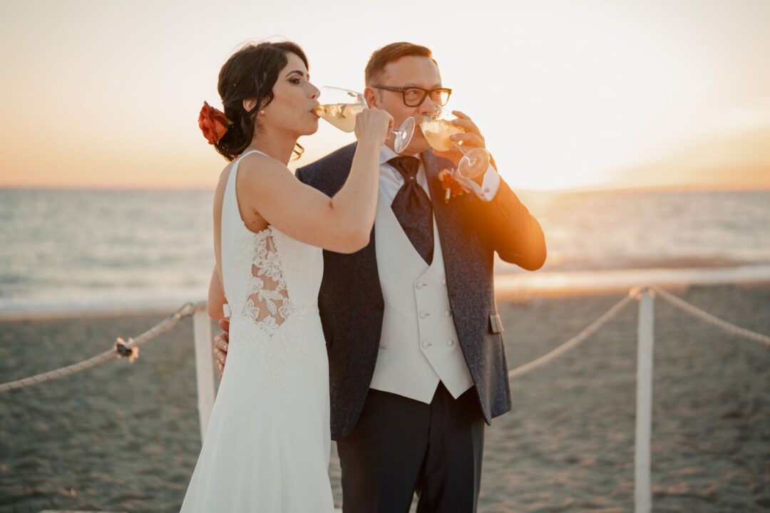 Newlyweds sharing champagne glasses at golden hour outdoors, a joyful crowd of wedding guests raising their glasses in a...