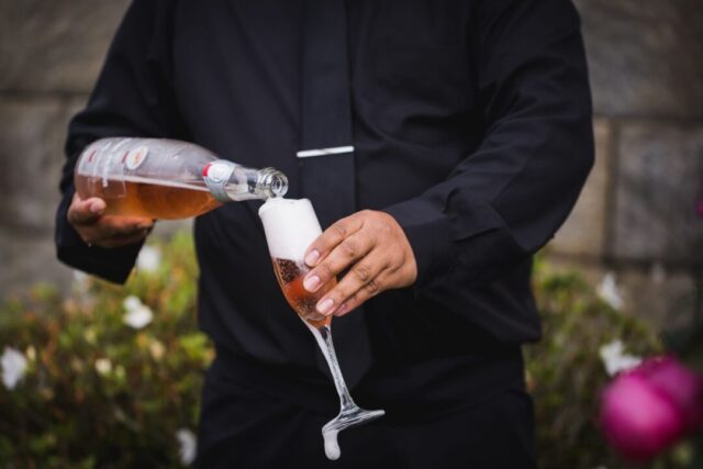 Bartender pouring a line of champagne flutes at an outdoor wedding reception, warm golden-hour lighting, elegantly...