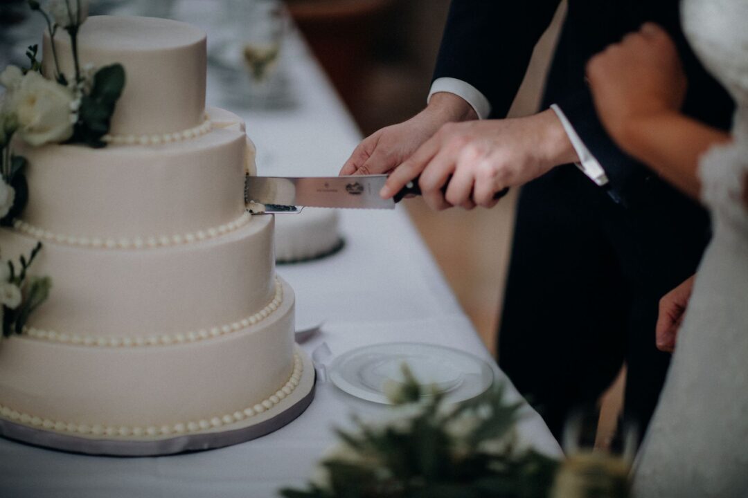Bride and groom cutting a three-tier white buttercream wedding cake decorated with fresh white peonies and trailing...