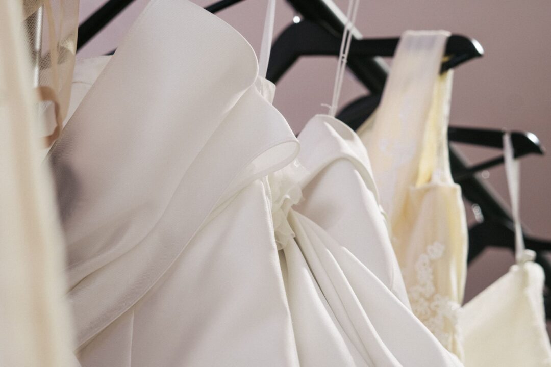 Neatly arranged display of white and ivory wedding gowns hanging on brass rails in a well-lit bridal boutique, showing a...