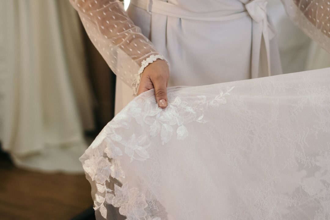 Close-up of a bride's hands delicately holding a sheer lace-edged bridal veil with a soft-focus ivory gown visible in the...