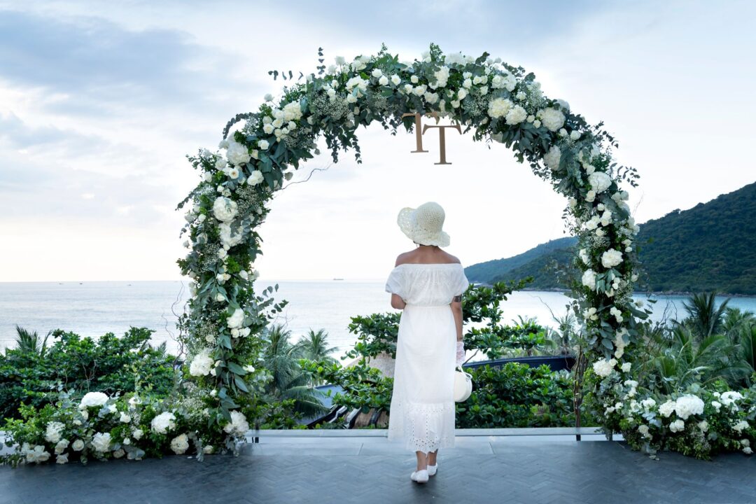 Professional wedding celebrant standing at a decorated outdoor ceremony arch, holding ceremony papers and smiling warmly...
