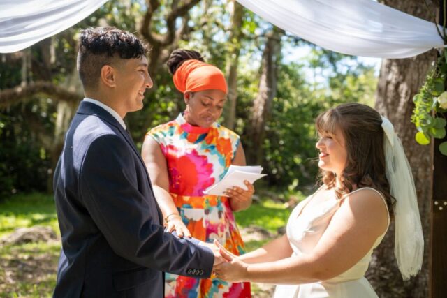 Couple exchanging vows with their wedding officiant at an outdoor ceremony, surrounded by seated guests in a garden setting