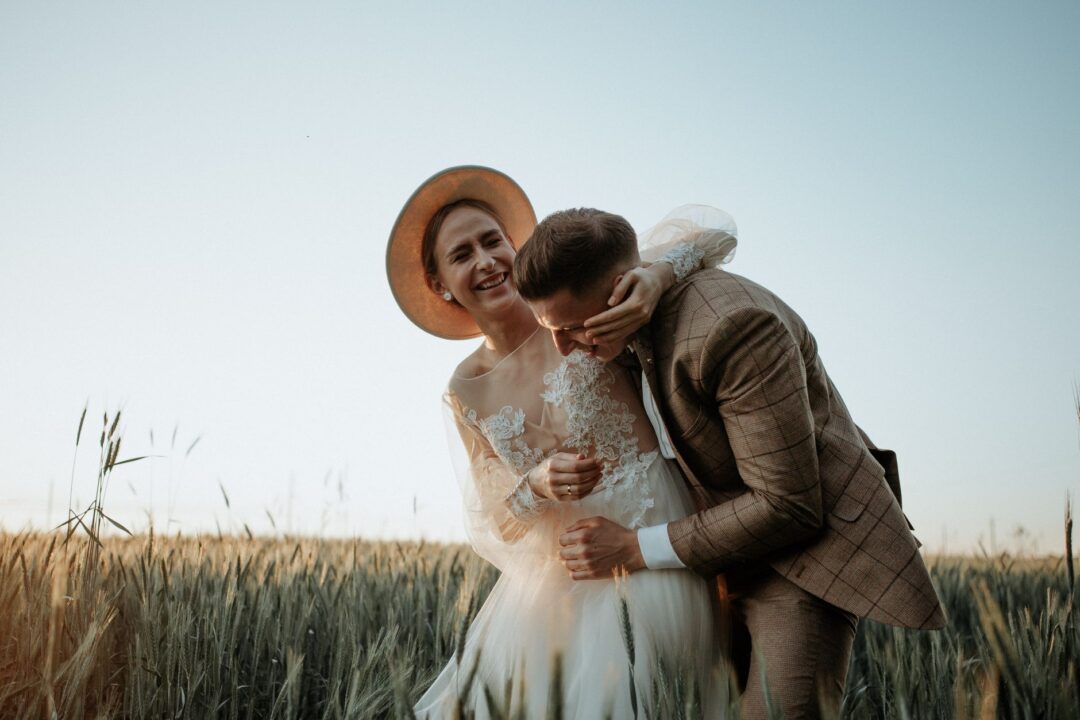Couple laughing together during a golden hour engagement session in a sunlit field, with the photographer crouching...