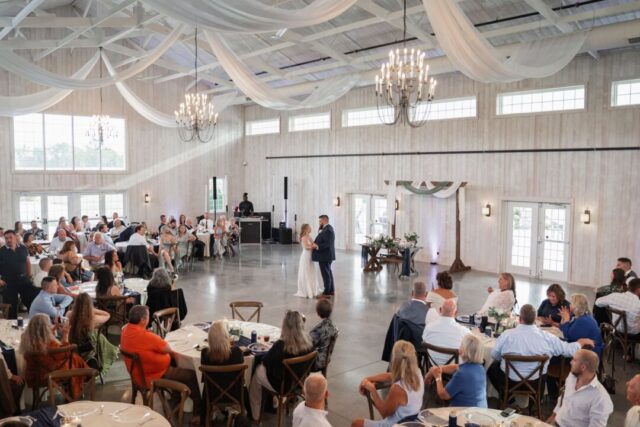 Wedding reception in full swing inside a light-filled barn venue, guests seated at round tables while the couple dances...