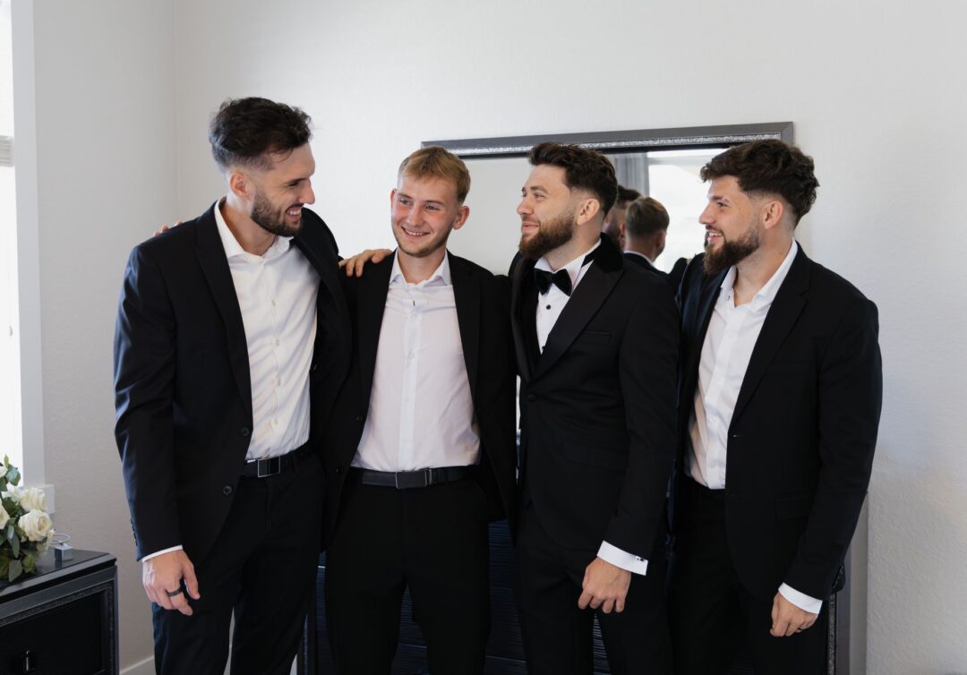 Four groomsmen in matching navy suits laughing together outside a sunlit venue entrance, boutonnieres pinned to their...