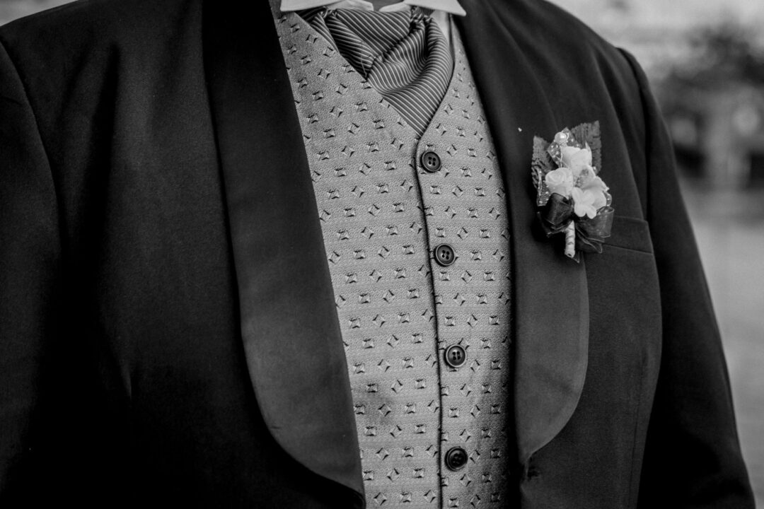 Close-up of a groom's lapel showing a white boutonniere pinned to charcoal wool, a folded cream pocket square tucked in...