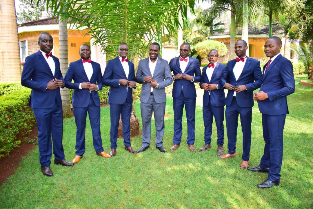 Groom and two groomsmen in coordinating charcoal suits walking together in bright afternoon sunlight on a wide city...
