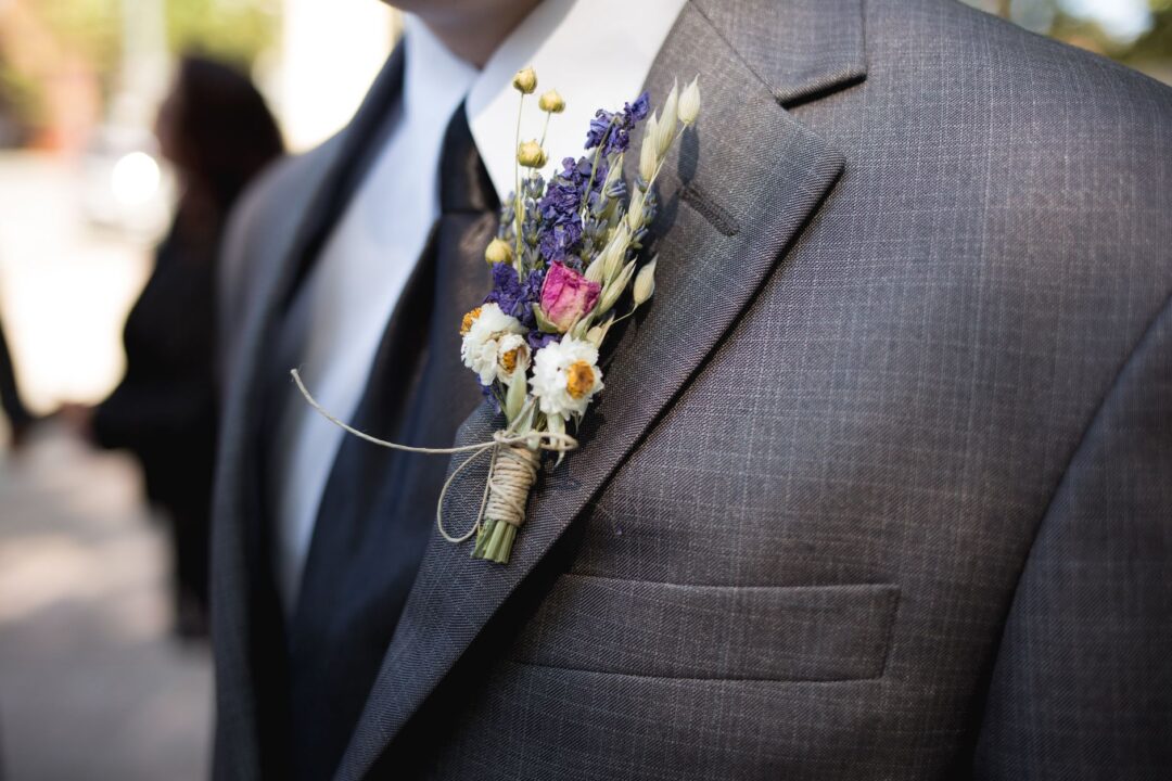 Groom in a charcoal wool suit standing in a bright hotel suite dressing room, adjusting his boutonniere in a full-length...