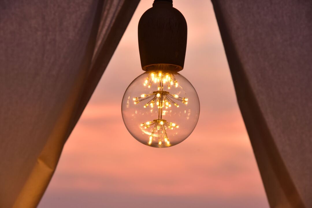 Close-up of sailcloth wedding tent fabric glowing with warm Edison bulb string lights at dusk, guests in formal attire...