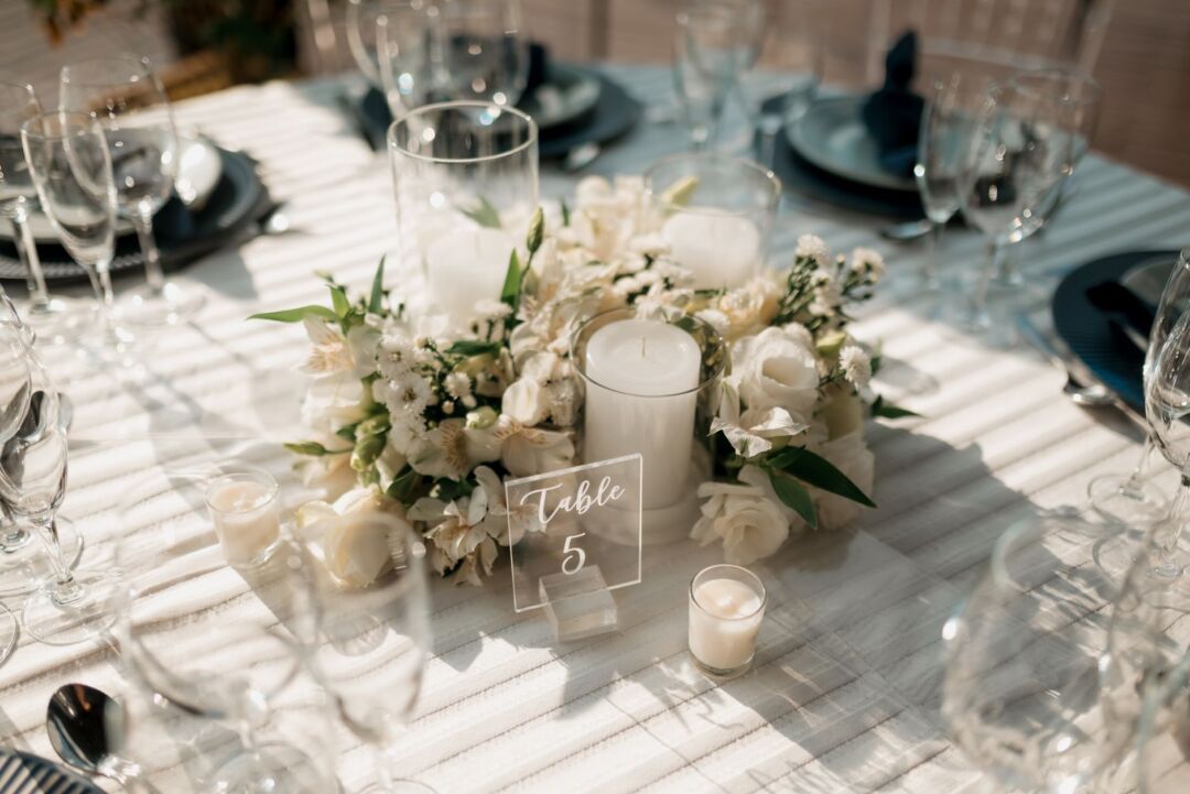 Round dining tables with white linen and floral centerpieces arranged inside a clear-sided frame tent, showing an...
