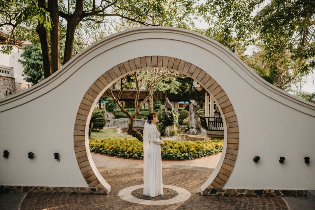 Bride standing outdoors at a garden ceremony venue seen from behind, wearing a two-tier chapel-length veil with a...