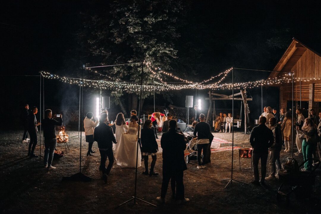 Rustic barn wedding reception with string lights, long wooden farm tables lined with greenery garlands, and warmly lit...