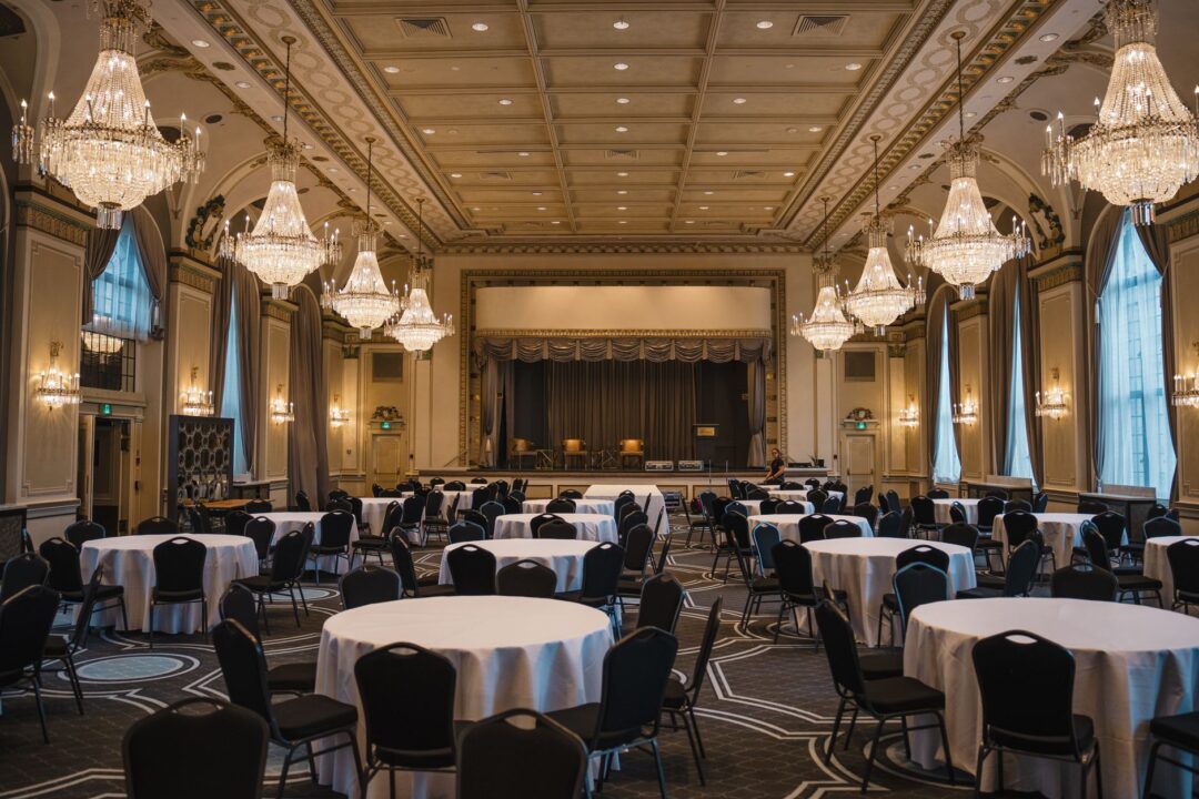 Grand hotel ballroom decorated for a wedding with round tables, white and gold table settings, tall floral centerpieces,...