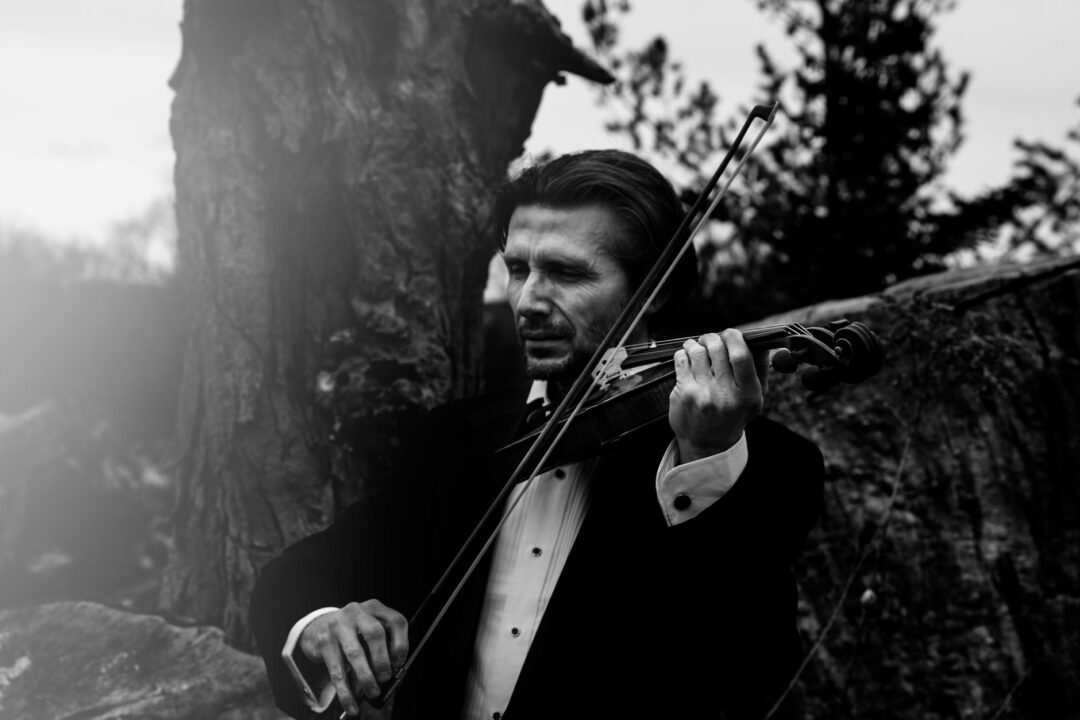 Close-up of a solo violinist performing at an intimate outdoor wedding ceremony, guests seated on wooden fold-out chairs...