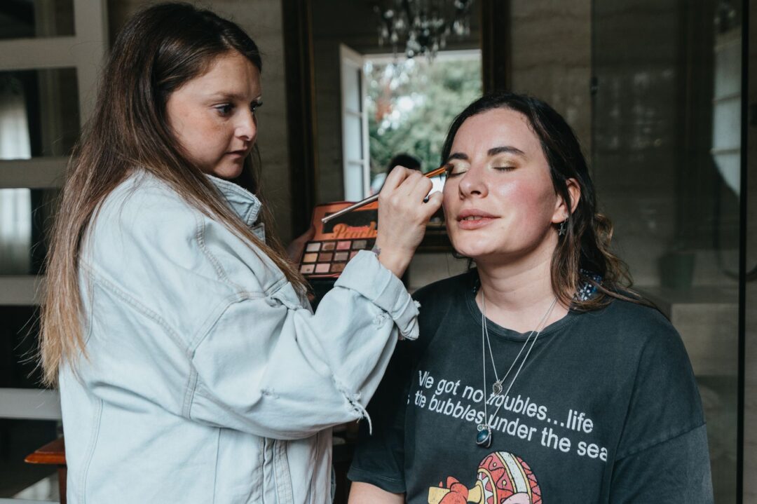 Professional makeup artist applying eyeshadow to a bride wearing a white robe, with a well-organized makeup kit open on...