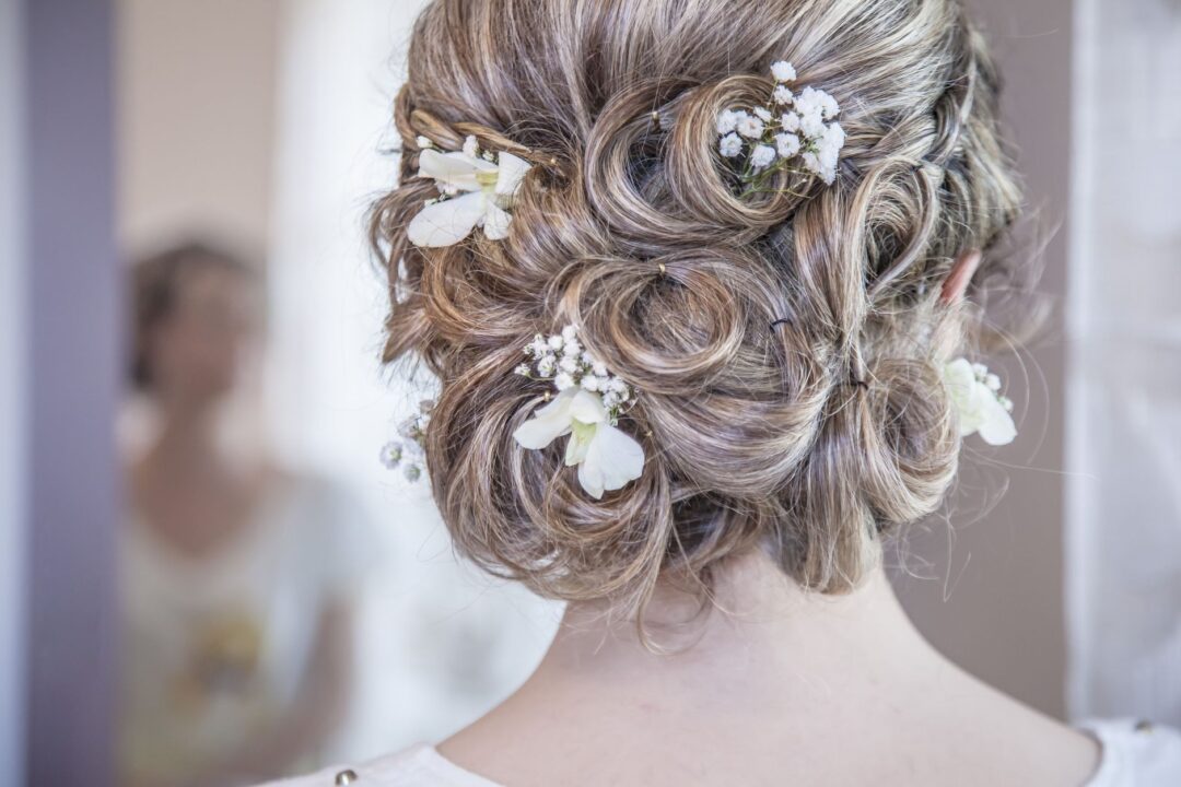 Bride's elegant updo photographed from behind, featuring soft curls pinned up with small white floral accents, with an...