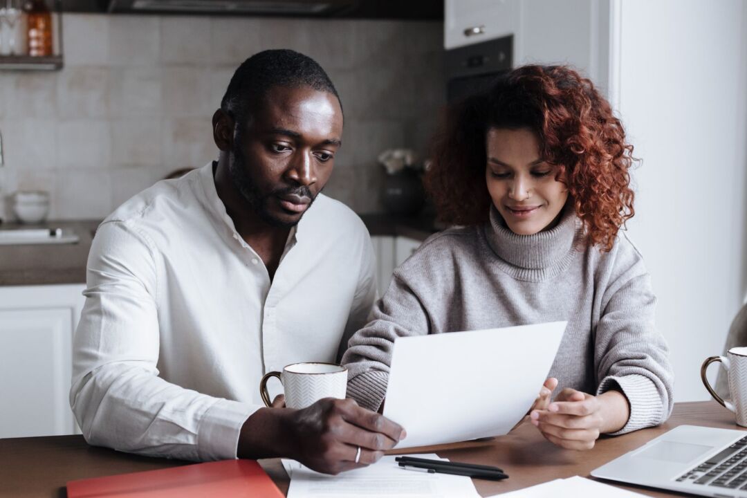 Couple sitting at a kitchen table together reviewing wedding planning documents, warm natural light, relaxed and focused
