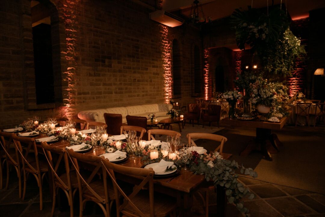 Romantic wedding reception with warm amber uplighting glowing along the walls of a hotel ballroom, bistro lights strung...