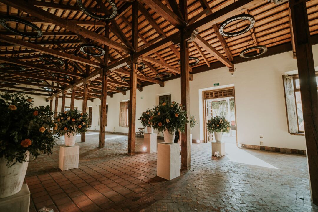 Scenic indoor wedding venue with exposed wooden beams, white fabric drapery, and long dining tables set for a reception,...