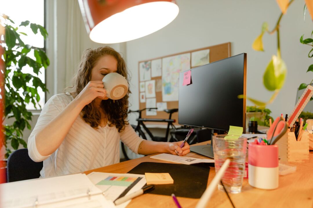 Woman with an engagement ring sitting at a kitchen table with a planner notebook and coffee mug, smiling as she writes,...