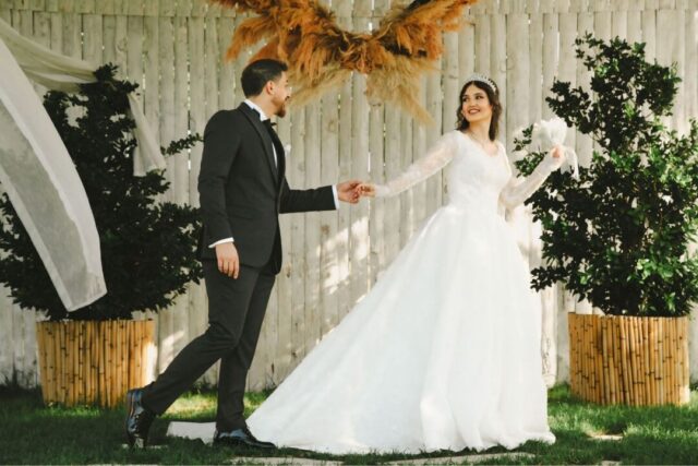 Couple in wedding attire holding hands and smiling at each other in a sunlit outdoor venue surrounded by autumn foliage...