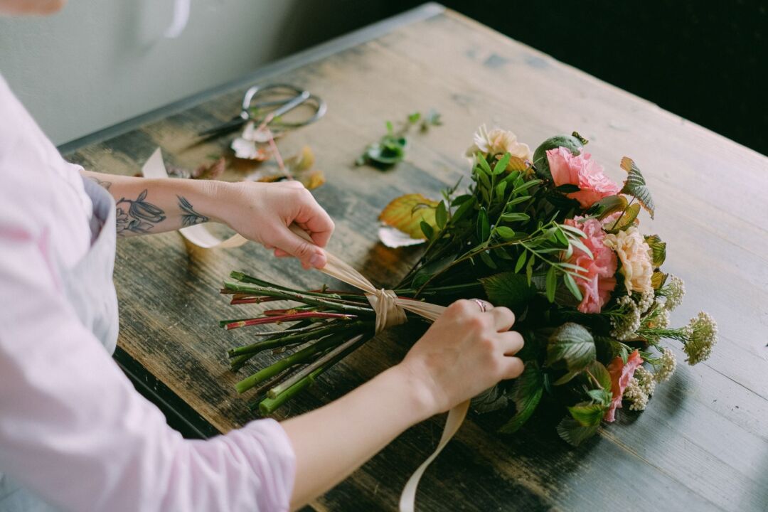 Close-up of a florist's hands arranging peony stems into a clear glass vessel on an outdoor table, surrounded by freshly...