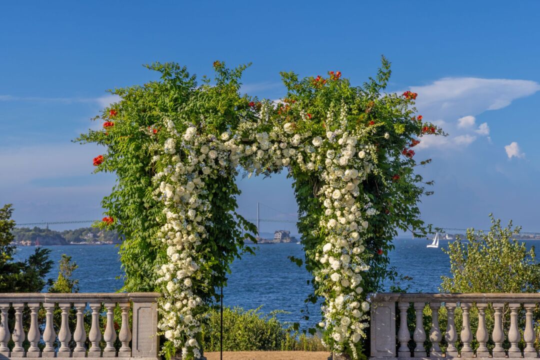 Outdoor summer wedding ceremony with a flower arch covered in garden roses, hydrangeas, and trailing greenery, warm...