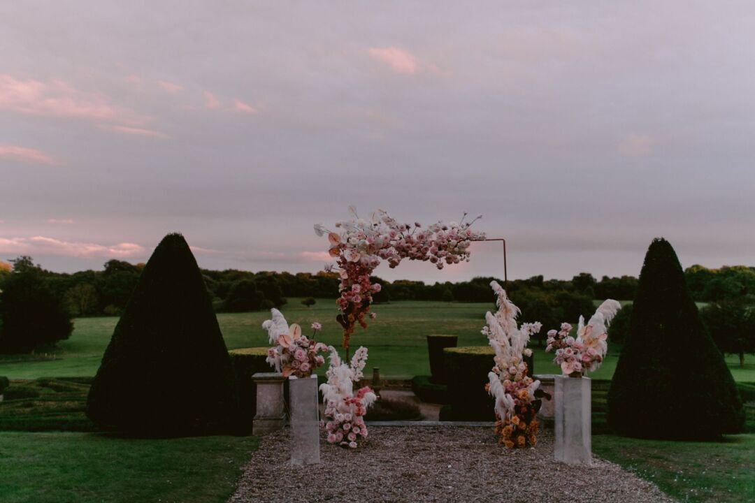 Lush wedding ceremony arch decorated with white peonies, garden roses, and trailing eucalyptus garlands, outdoor garden...