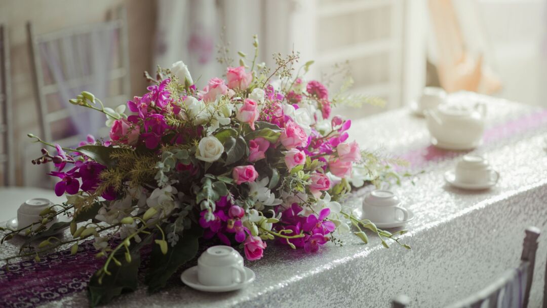 Styled wedding reception table with fresh flowers in small bud vases and a lush artificial greenery garland runner, warm...