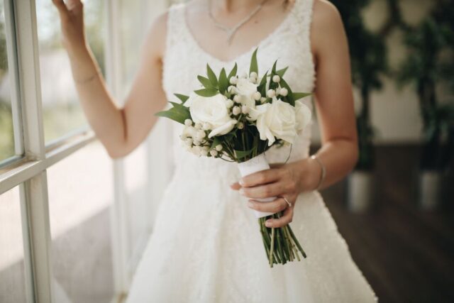 Bride in soft natural light holding a lush bouquet of white peonies and garden roses, close-up documentary style,...