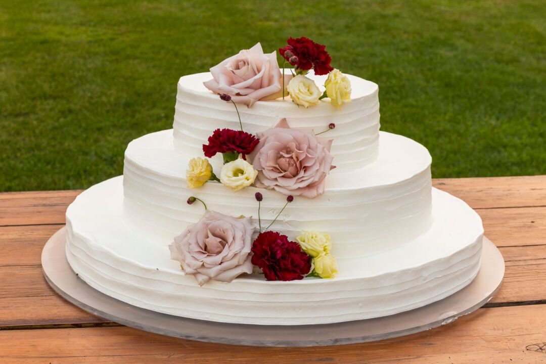 Three-tier wedding cake with white textured buttercream and gold leaf accents displayed on a marble cake stand surrounded...