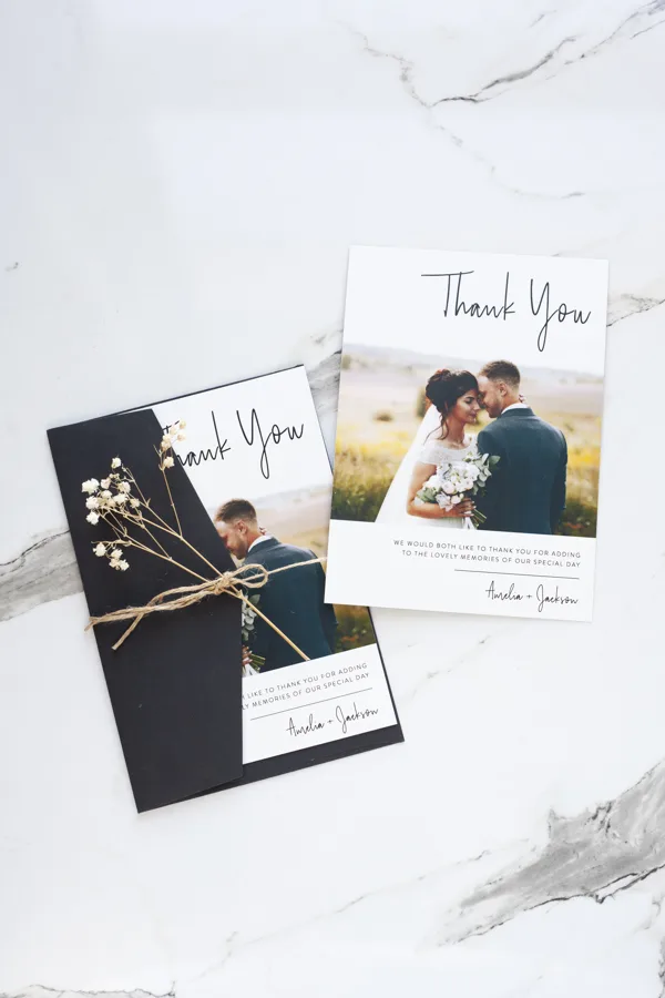Paperlust Linear wedding thank you card with script thank you, couple photo and a short pre-printed message, styled on marble with a black envelope and dried flowers