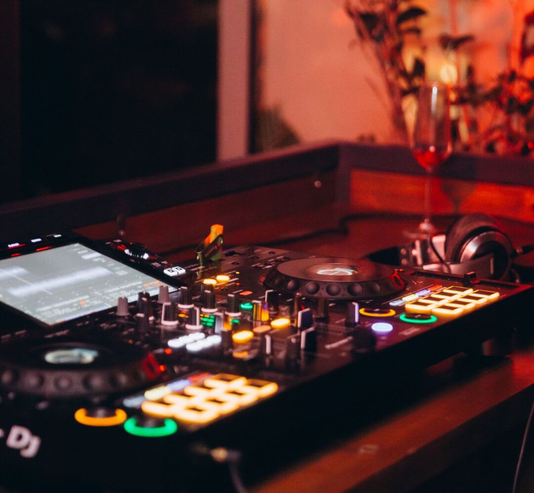 Professional wedding DJ behind a lit console at a modern reception venue, mixing equipment glowing, guests on the dance...