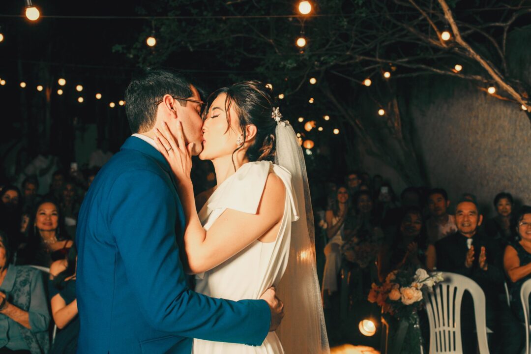 Joyful couple sharing their first kiss as newlyweds at an outdoor ceremony, guests cheering and rising from their seats...