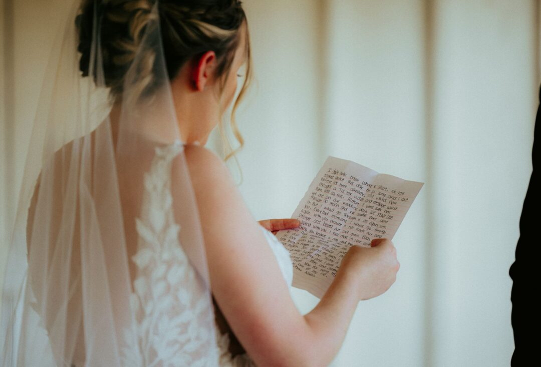 Bride at the altar reading her handwritten vows from a small card, a videographer with a cinema camera visible in soft...