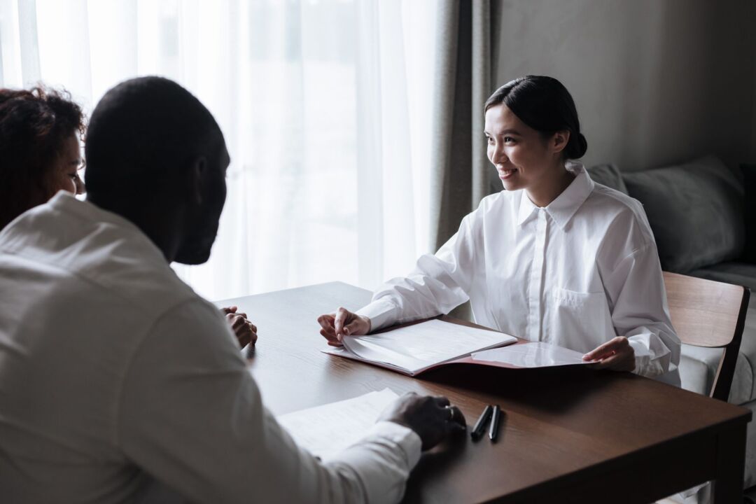 Couple sitting across a table from a wedding vendor reviewing printed documents together in a bright modern office,...