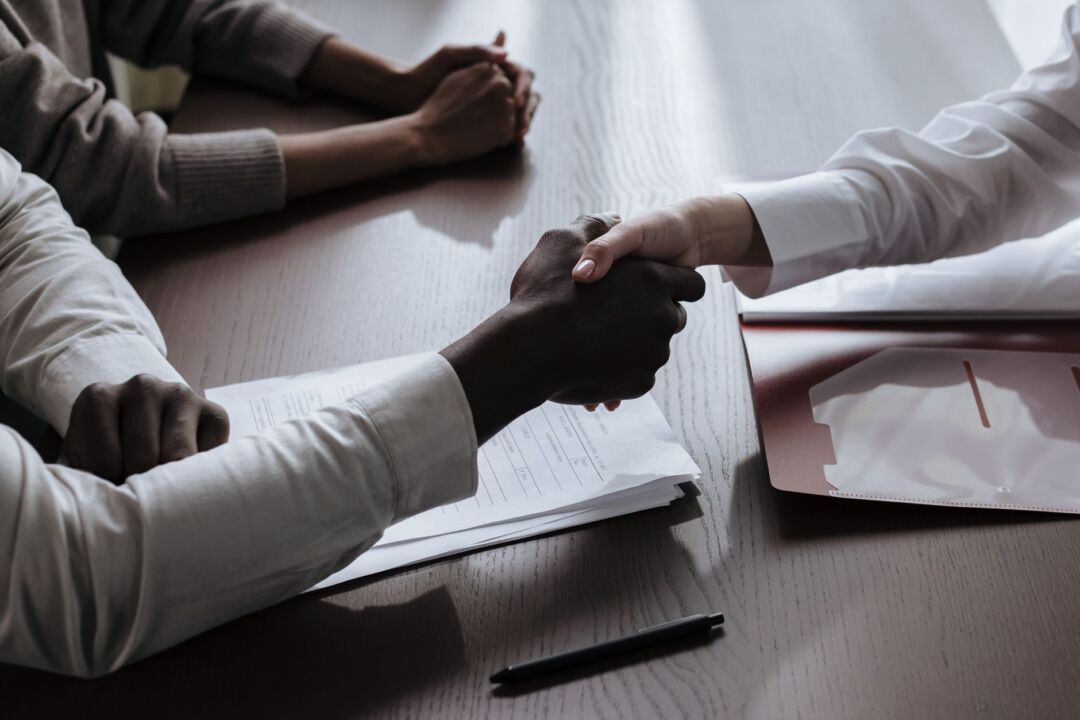 Close-up of two people shaking hands across a desk after finalizing an agreement, an open folder of documents and a pen...
