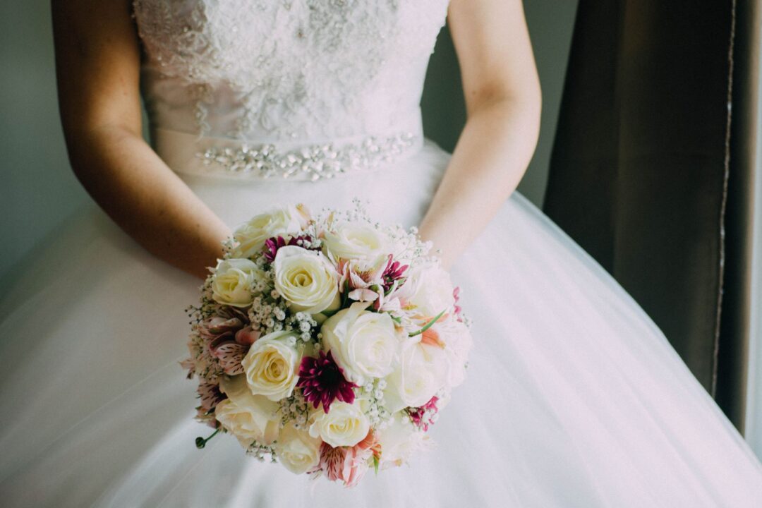 Relaxed bride in her wedding gown holding a bouquet and laughing with her maid of honor while a coordinator adjusts a...