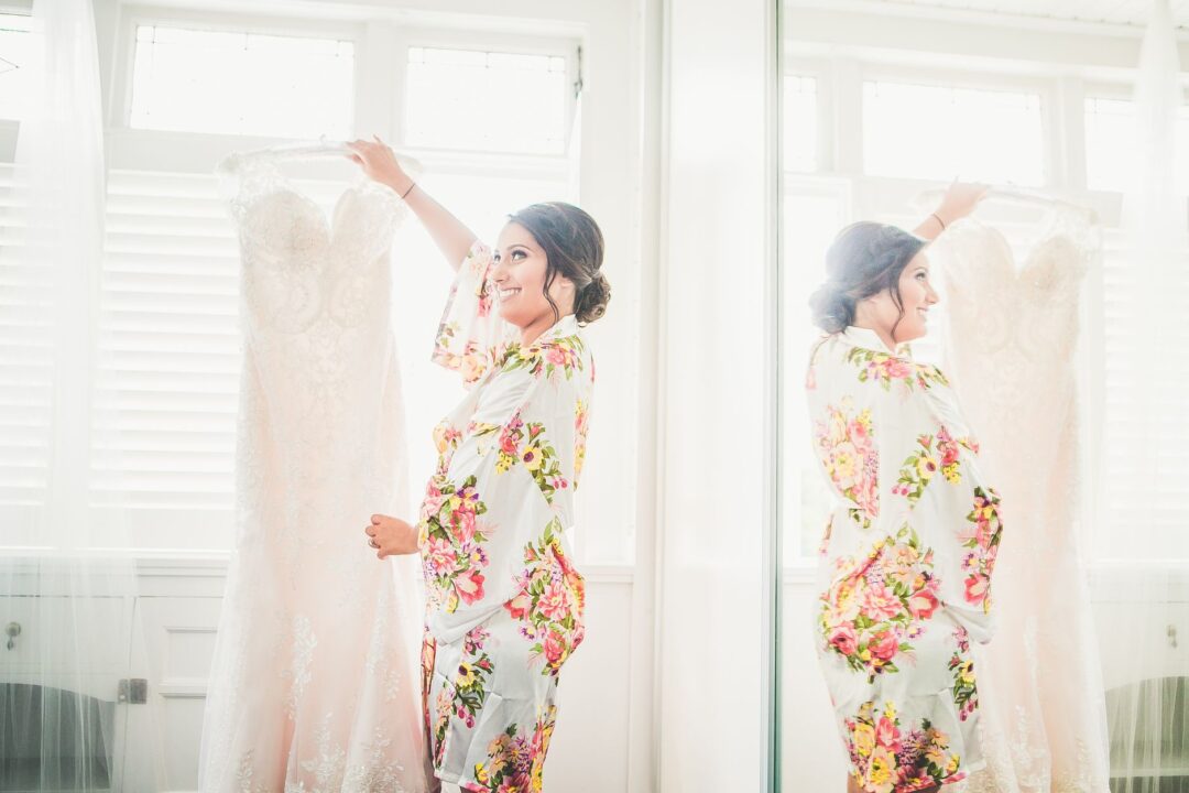 Bride standing near a large window in soft natural daylight, photographing her trial look in a mirror to document the...