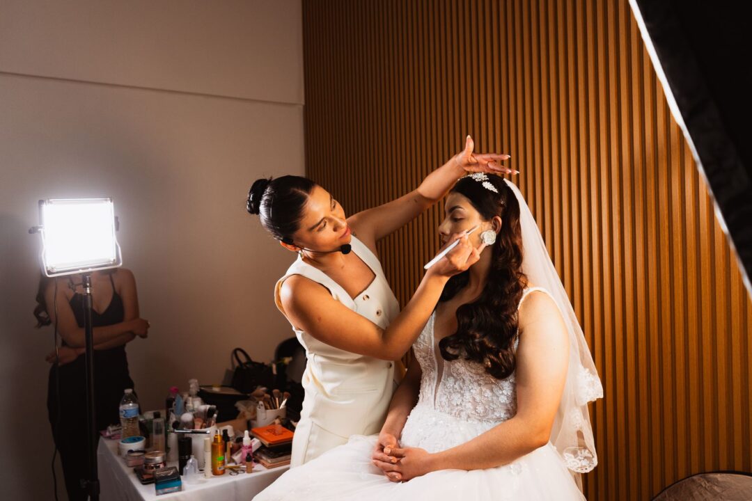 Makeup artist (or hair stylist) WORKING on a bride, applying makeup at a vanity/mirror, a hair-and-makeup trial in progress