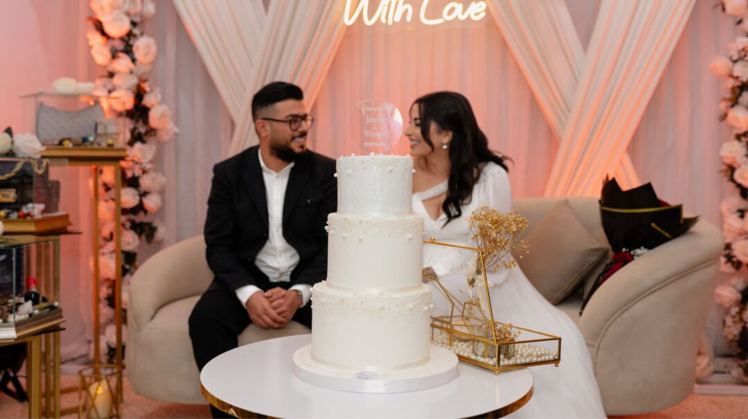 Couple seated side by side at a bakery consultation table, looking at a cake design portfolio together, with handwritten...