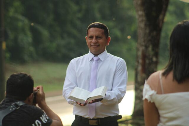 Smiling couple stands before their officiant in an airy outdoor venue, surrounded by greenery, as they exchange vows on a...