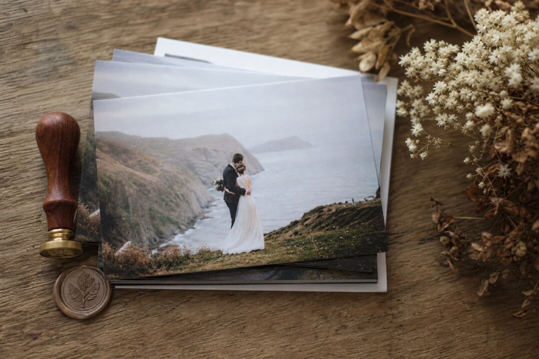 A flat-lay of printed elopement announcement cards stacked on a wooden table with a wax seal stamp and dried flowers beside them.