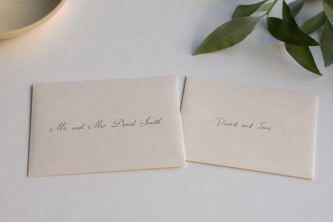 Two cream-colored place cards with handwritten names sit on a white surface next to fresh green leaves and a ceramic bowl.